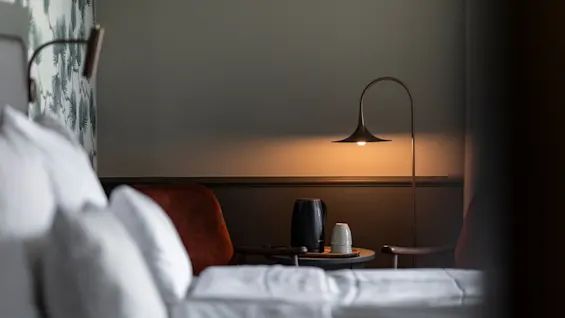 Mukava makuuhuoneen näkymä, jossa sänky etualalla, lamppu valaisee sivupöytää vedenkeittimellä ja kupeilla.