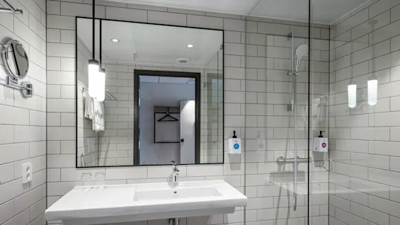 Vask reflekteres i et badeværelse spejl, omgivet af hvide flisevægge; en glasbruser med regnbruserhoved er synlig til højre, og toiletartikler er monteret på væggen.