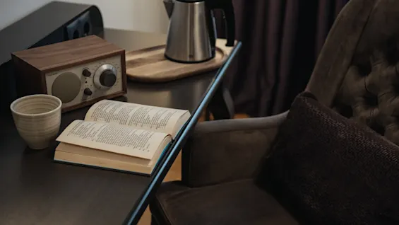 Viihtyisä lukunurkkaus, jossa avoin kirja, kuppi, radio ja vedenkeitin tummalla työpöydällä mukavan nojatuolin vieressä.