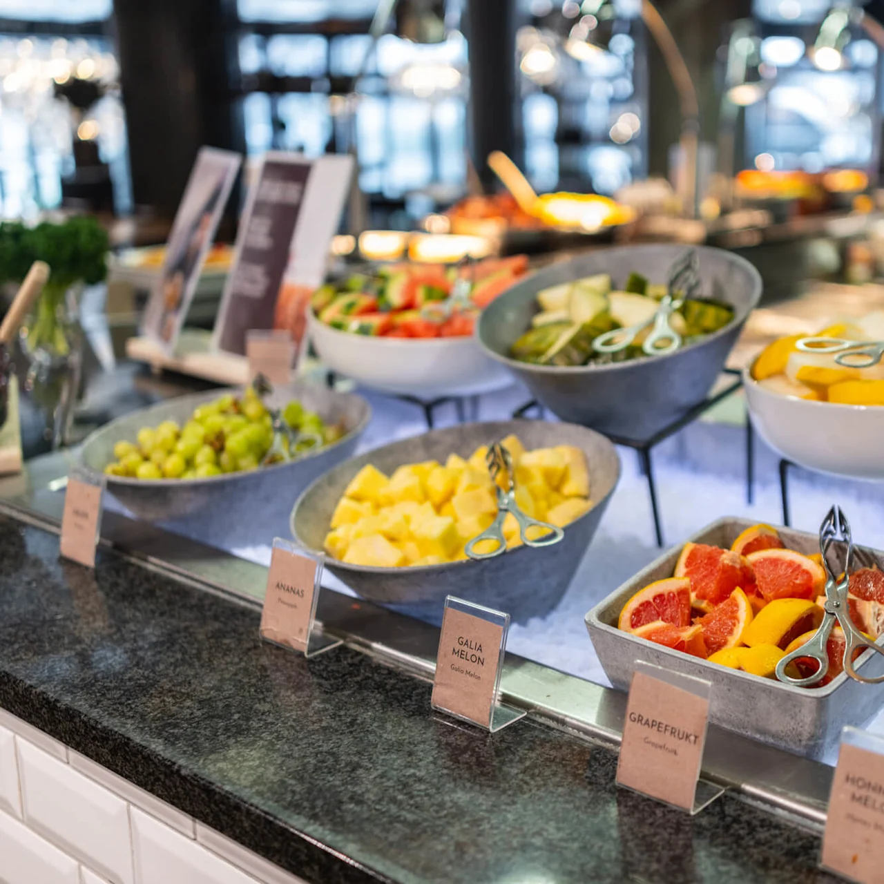 Et buffébord viser forskjellige oppskårne frukter i boller med serveringstenger, plassert på is. Nærliggende etiketter lyder: "Honningmelon", "Grapefrukt", "Galiamelon", "Ananas", "Vindruer".