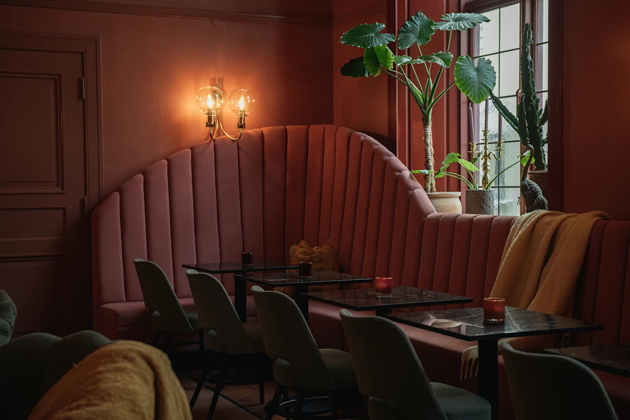 Røde polstrede siddepladser kurver sig langs cafévæggen under varm belysning, med grønne planter på vindueskarmen og små borde med grå stole, der skaber en hyggelig, indbydende atmosfære.