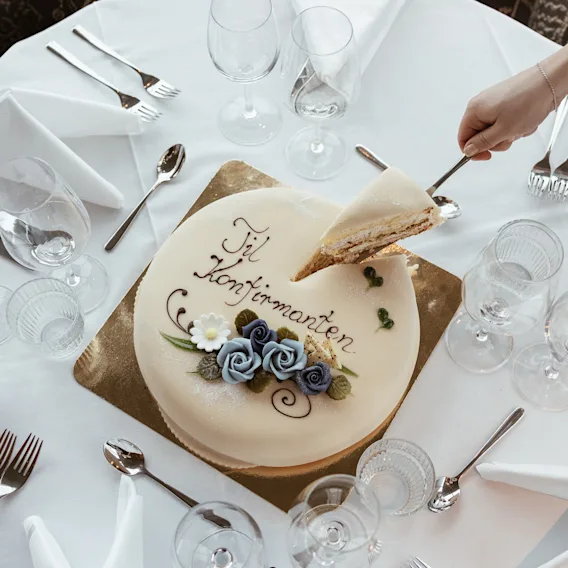 En hånd serverer et stykke hvit festkake dekorert med blå blomster og teksten Til Konfirmanten på et dekket bord.