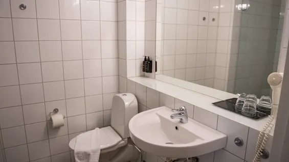 Toilet with a towel on the seat, adjacent to a sink and mirror in a tiled bathroom featuring toiletries, glasses, and a wall-mounted hairdryer.