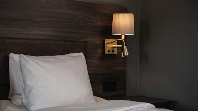 Bed with white pillow and bedding, situated beneath a wooden headboard. A wall-mounted lamp with a beige shade illuminates the setting, creating a cozy bedroom atmosphere.