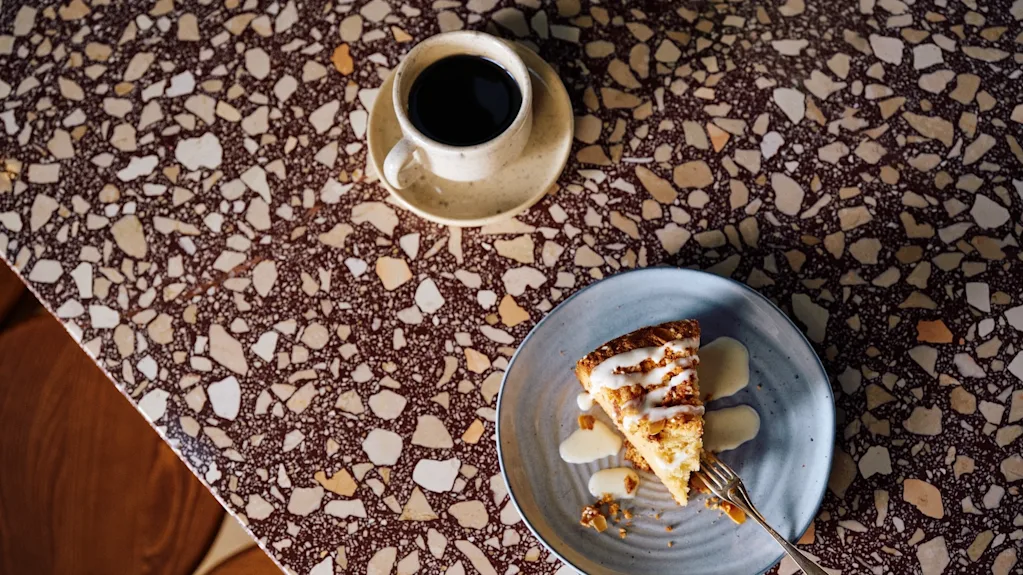 Et stykke kage med glasur ligger på en blå tallerken med en gaffel, placeret på et mønstret stengulv ved siden af en kop sort kaffe.