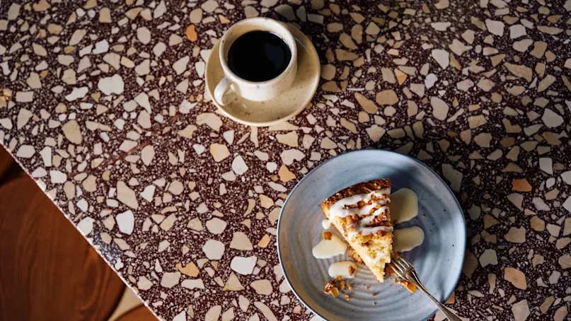 A slice of cake drizzled with icing sits on a blue plate with a fork, placed on a patterned stone table next to a cup of black coffee.