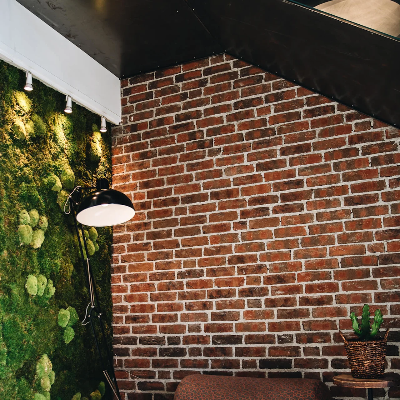 Floor lamp illuminating a cozy seating area with a gray chair, next to a green moss wall. Brick walls form the background, with a small table holding a potted cactus.
