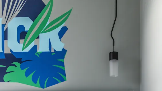 A bed with white sheets sits against a dark headboard; a pendant light hangs above. Wall decor shows the text "ICK" with green leaves.