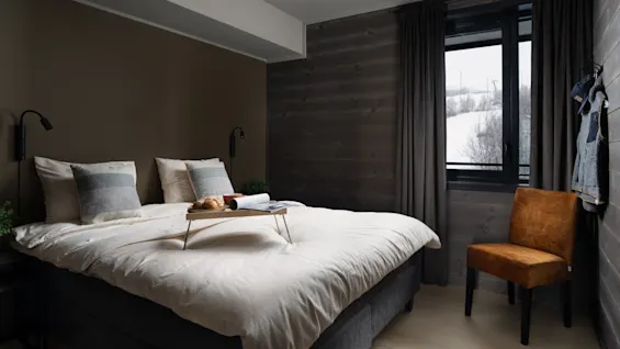 Seng med morgenmadsbakke står i et hyggeligt, moderne soveværelse. En brun stol står ved siden af vinduet, der afslører et sneklædt landskab. Mørke gardiner indrammer udsigten, og der hænger tøj på væggen.
