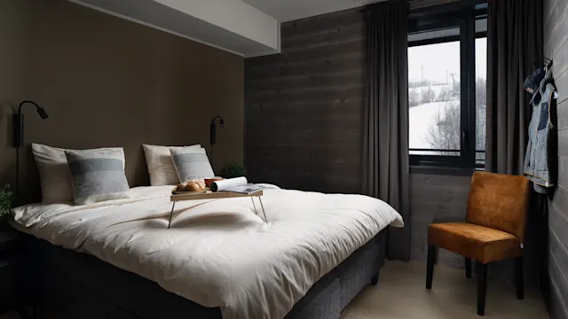 Bed with breakfast tray sits in a cozy, modern bedroom. A brown chair is beside the window, revealing a snowy landscape. Dark curtains frame the view, and clothes hang on the wall.