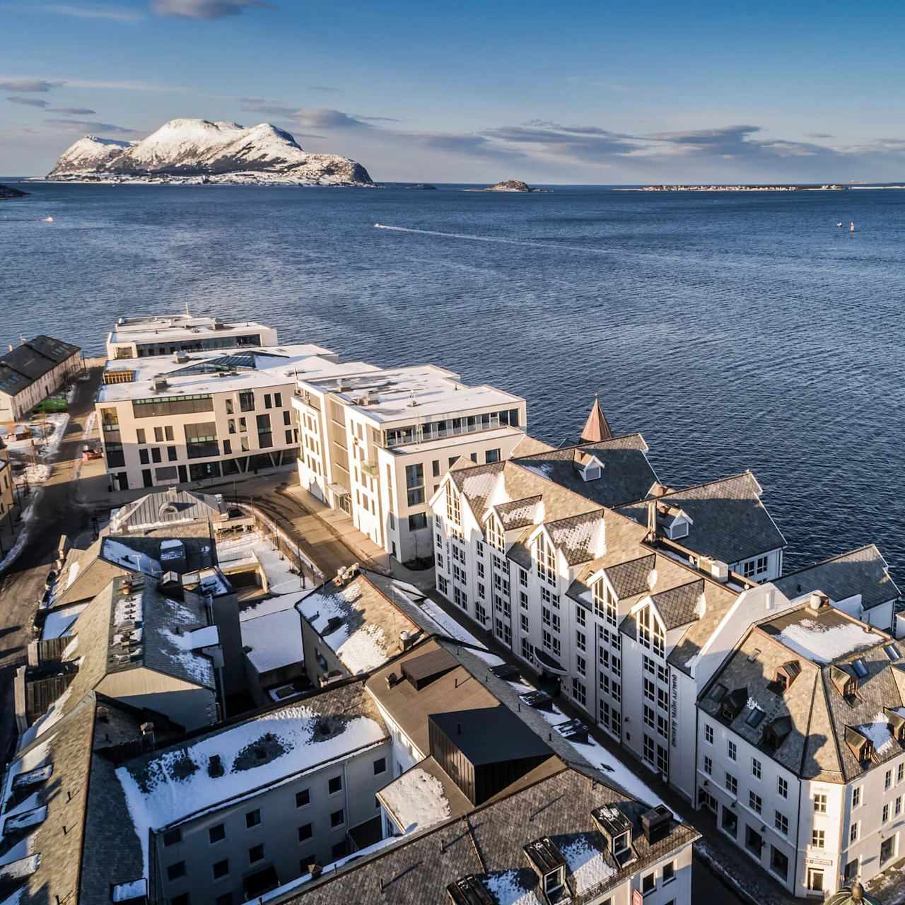 Snefækkede bygninger ligger langs en kyst, der vender ud mod et stort vandområde. I det fjerne rejser snedækkede bjerge sig under en klar blå himmel.