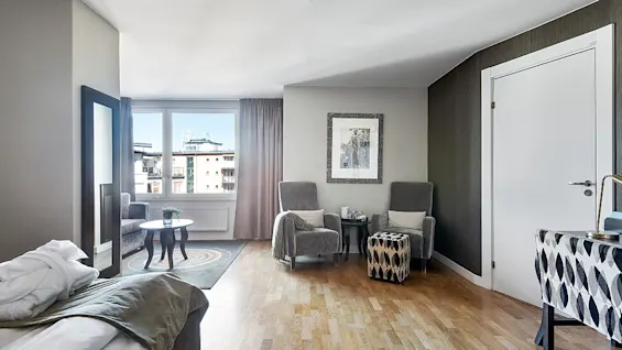 A well-lit, modern bedroom with a large bed, mirror, seating area, and balcony doors, showcasing an urban view.