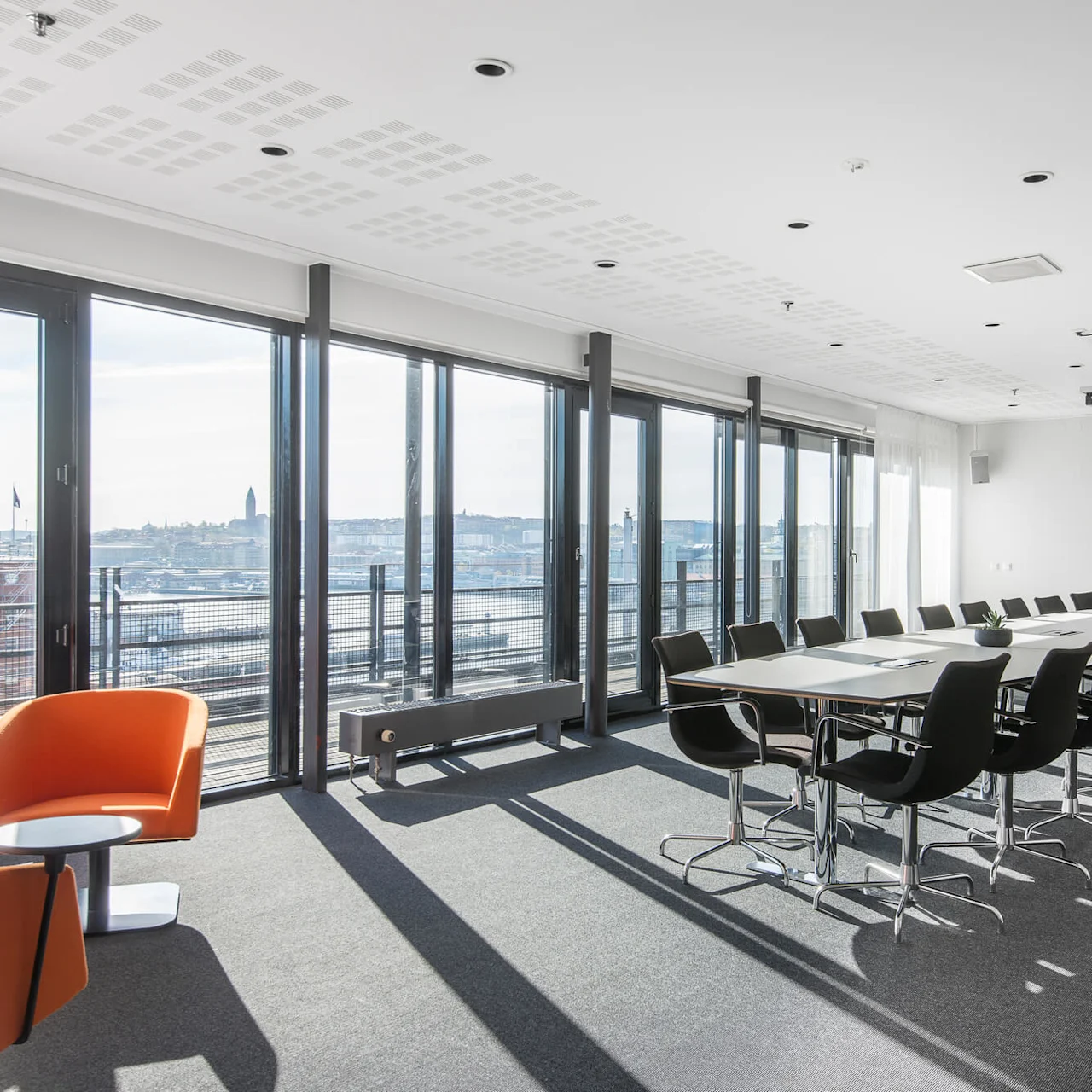 A conference room with a long table and black chairs, facing large windows with a city view. Two orange chairs and small tables are in the corner.
