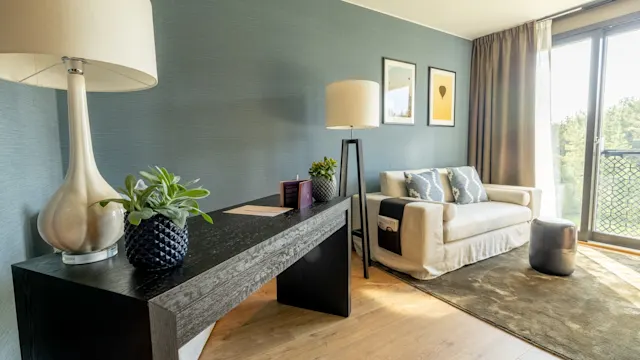 Et moderne stue med et skrivebord med planter og en lampe, ved siden av en sofa med puter. Innrammet kunst pryder blå vegger, mens sollys strømmer gjennom store vinduer.