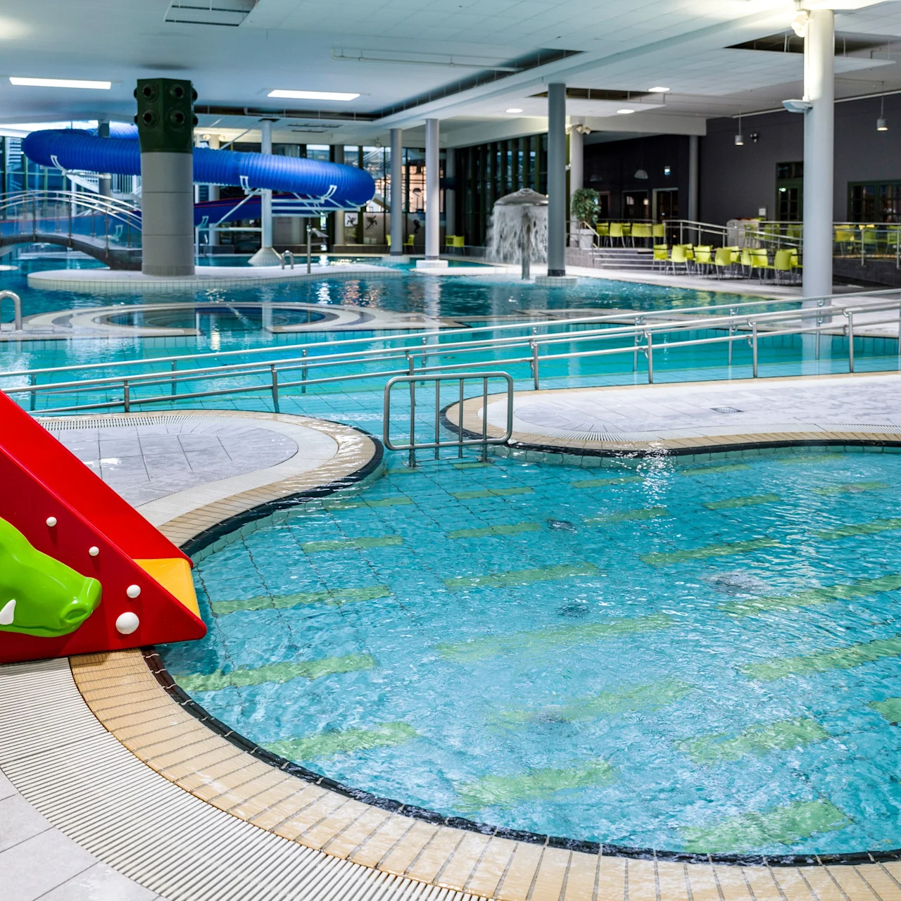 A children's red slide, shaped like a friendly animal, descends into a shallow indoor swimming pool area. Surrounding features include a larger pool with a blue water slide and seating.