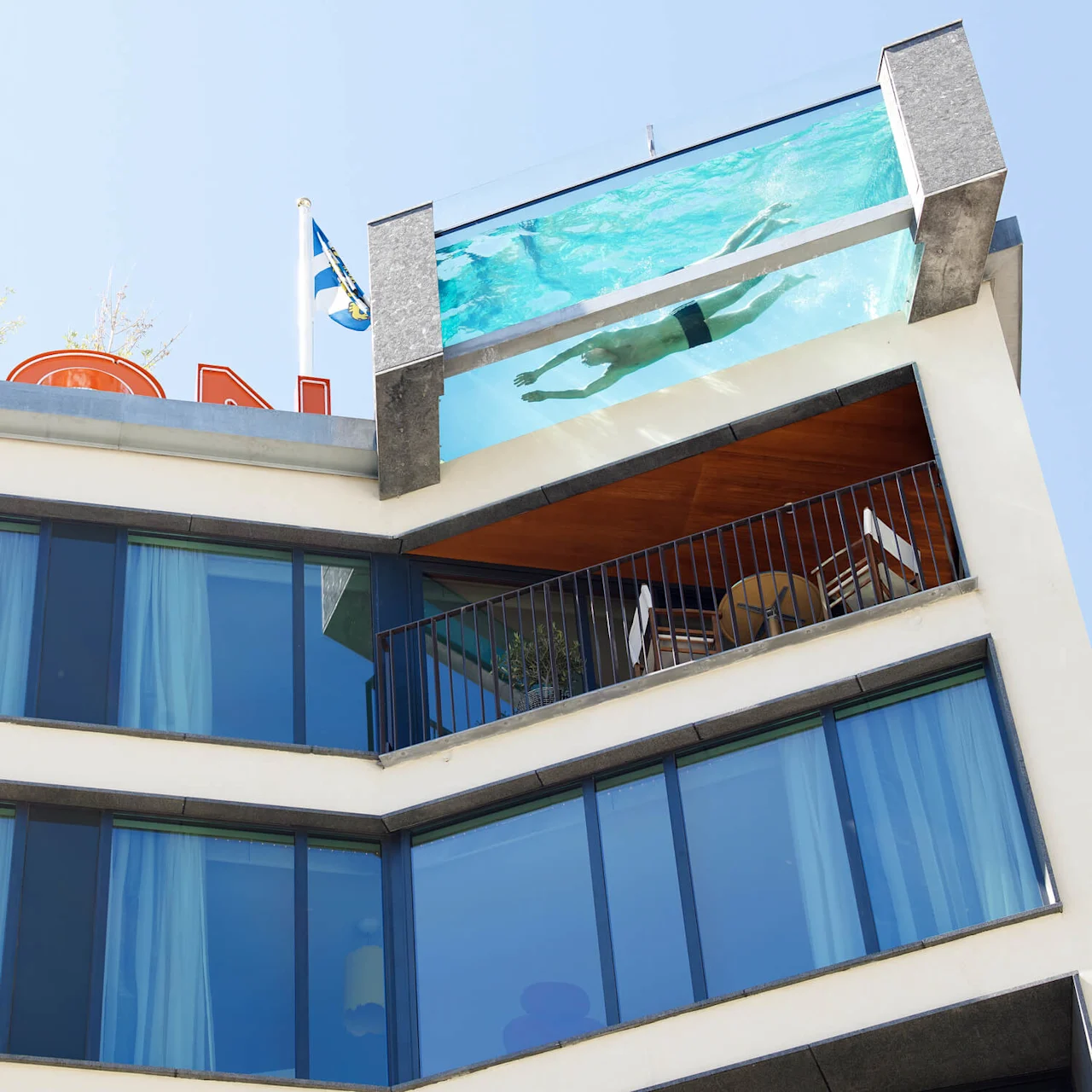 A rooftop pool with a swimmer above a modern building facade, featuring glass windows and a balcony with seating. The sky is clear. Text: "KAUAI PALACE HOTEL."