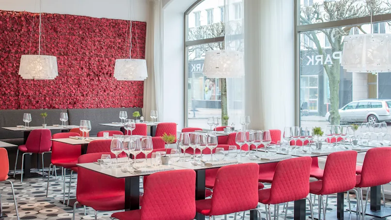 Elegant dining tables and red chairs are set with glassware in a modern restaurant. Large windows provide a view of the street outside. A textured red wall adds to the decor.