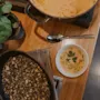 En gryta med krämig soppa står på ett träbord bredvid en skål med frön, med en växt i närheten och en sked och liten soppskål i sikte.