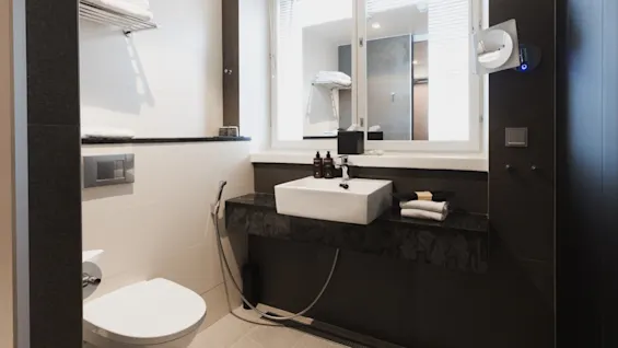 Et moderne badeværelse har et hvidt toilet og håndvask med toiletartikler, herunder flasker mærket "hair" og "body." Håndklæder ligger på hylder; sollys strømmer gennem et stort vindue med persienner.