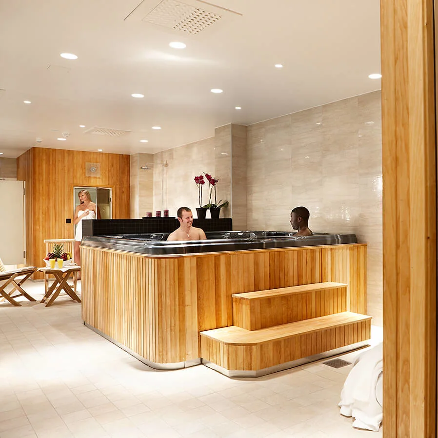 Hot tub with two people relaxing indoors, surrounded by wooden and tiled walls, next to a sauna with a glass door. A person stands near a table with towels.