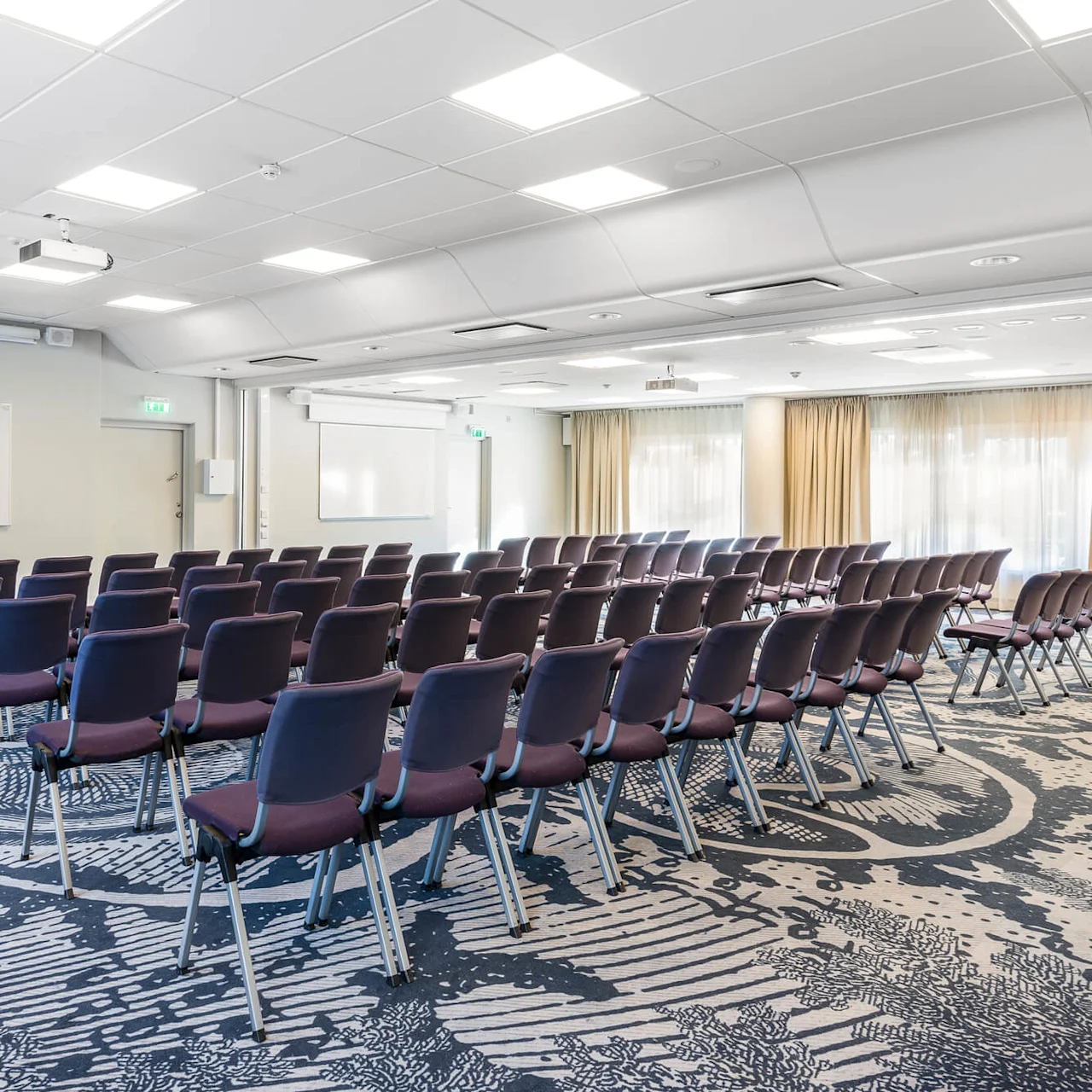 Tomma rader med stolar vetter mot två whiteboards i ett ljust konferensrum med mönstrad matta och taklampor. Beige gardiner täcker fönstren på den högra väggen.