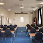 Et rummeligt konferencelokale med rækker af borde og stole, et projektorlærred og en whiteboardtavle, klar til en præsentation.