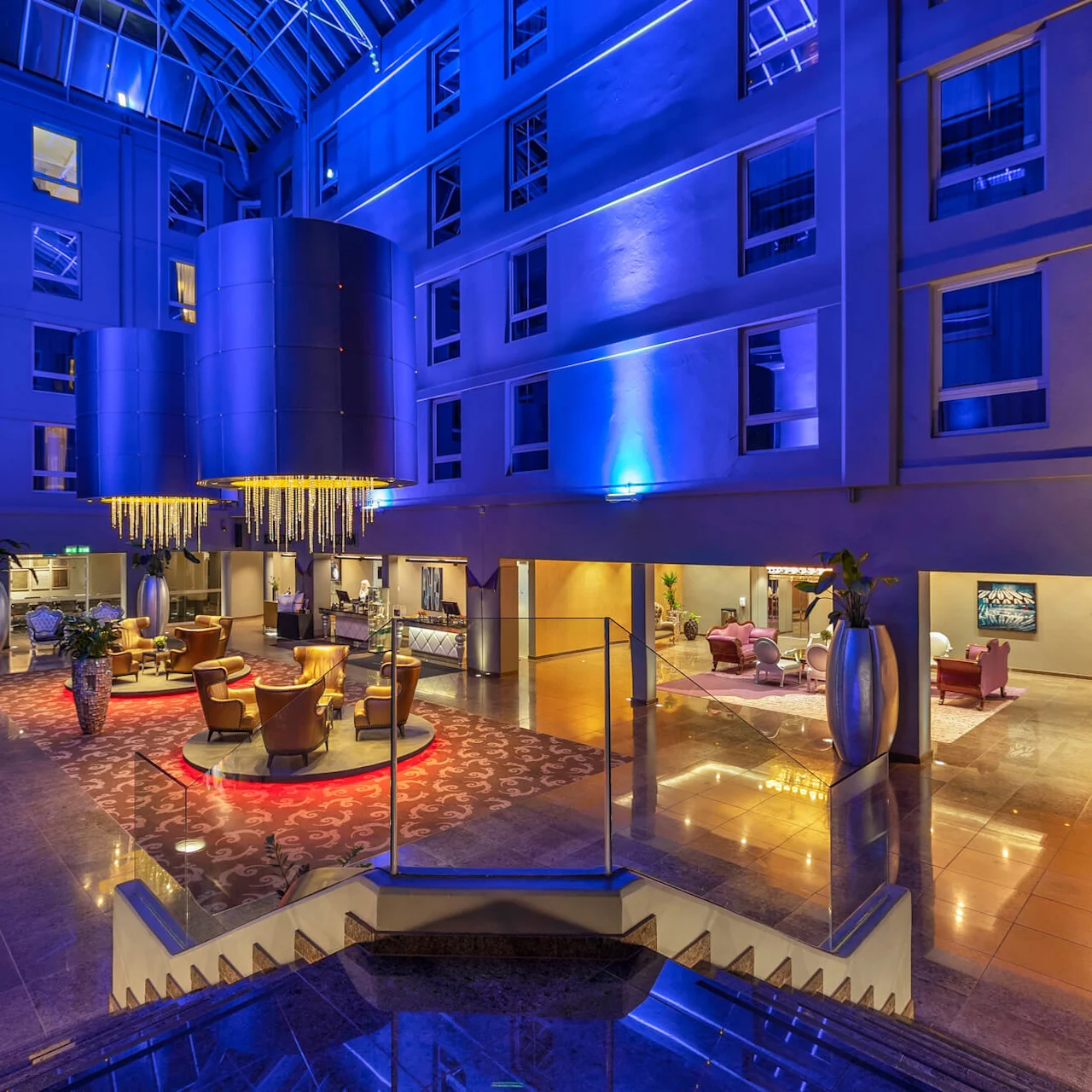 Et stort atrium med moderne lysekroner oplyser en hotellobby. Varm belysning fremhæver siddeområder, omgivet af høje planter. Blå accentbelysning forbedrer stemningen i det fleretagers rum.
