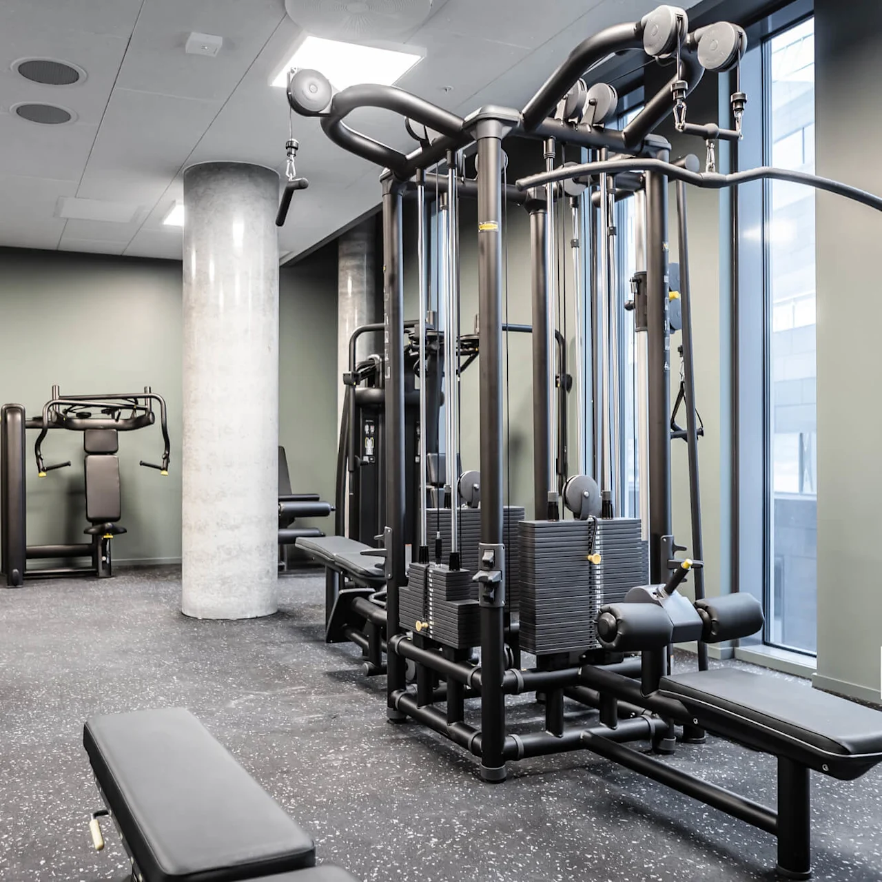 Workout machines are positioned in a bright, modern gym. Cable machines and benches are arranged around a central pillar, facing windows that allow natural light to illuminate the space.