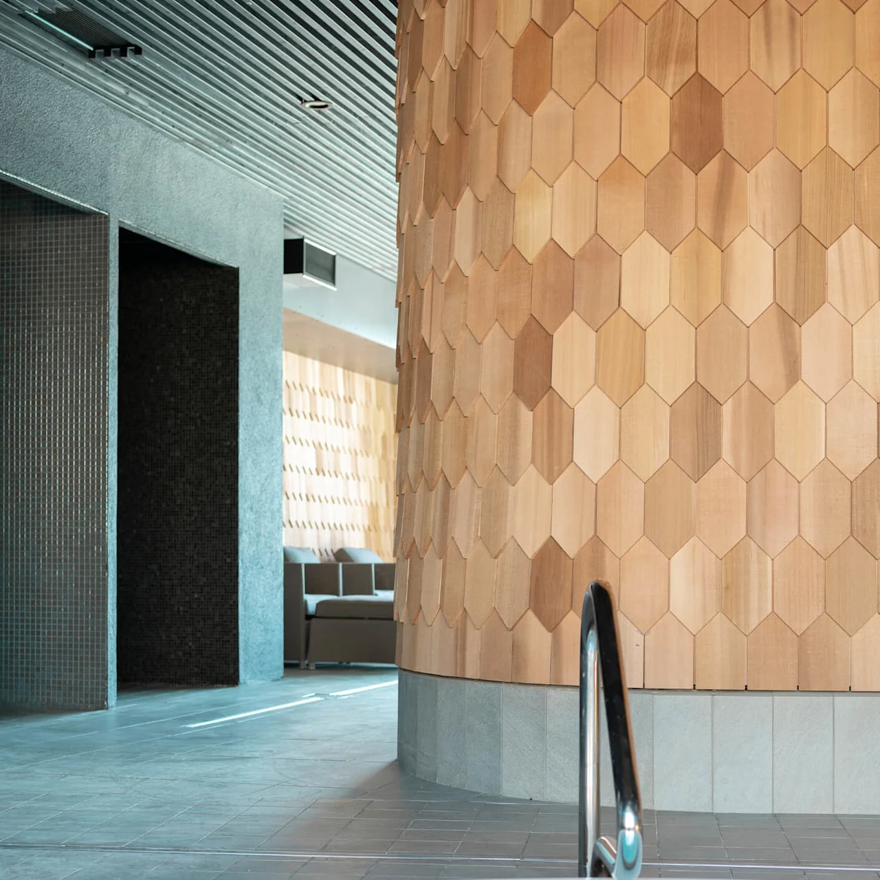 Hot tub with a metal handrail sits in a sleek, modern spa interior featuring hexagonal wood paneling and grey tiled walls, under a slatted ceiling with ambient lighting.