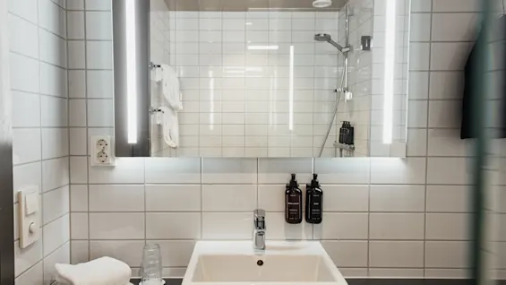 Valoisa, moderni kylpyhuone, jossa on valkoinen pesuallas, valaistu peili ja kaakeloitu suihkutila hygieniatuotteineen.
