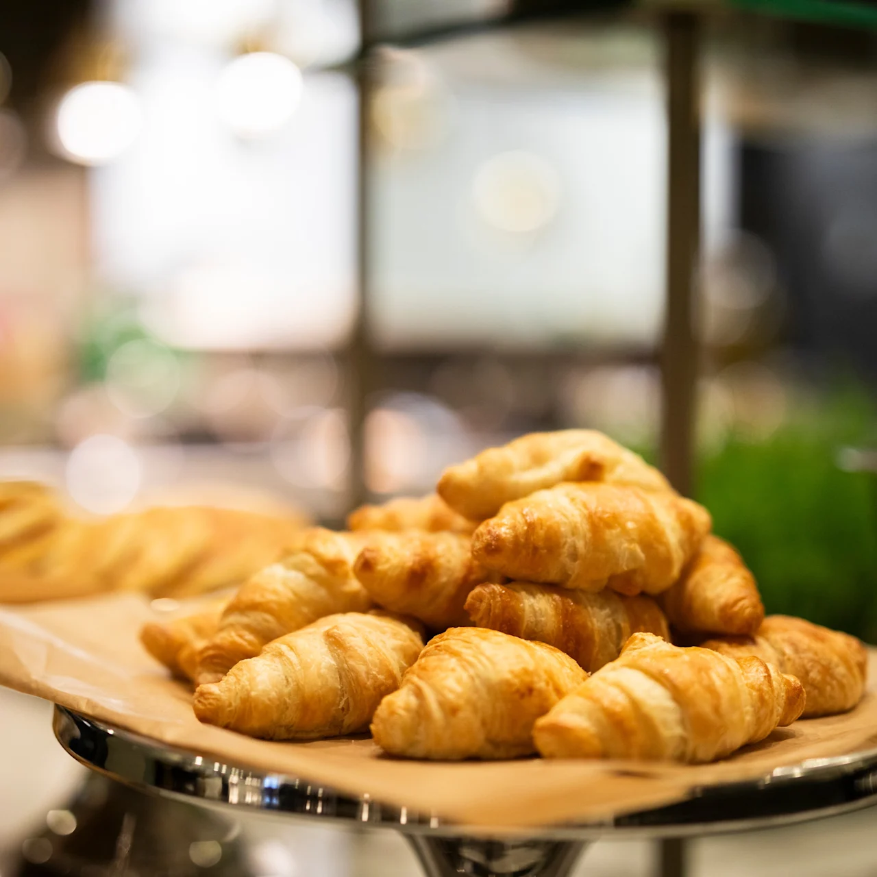 Golden croissants rest on a paper-lined tray, displayed prominently. The setting appears to be a bakery or a breakfast buffet with a blurred, warm-lit background.