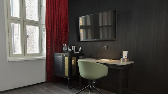A neatly organized office corner with a desk, chair, and a mounted television, in a room with dark walls and a window.
