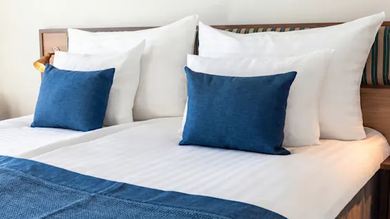 A neatly made hotel bed with crisp white sheets, blue decorative pillows, and a blue throw. Quality Hotel The Book.