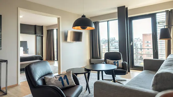 A modern living room features black chairs, a gray sofa, and a round table under a pendant light. Large windows display a cityscape. A bedroom is visible in the background.
