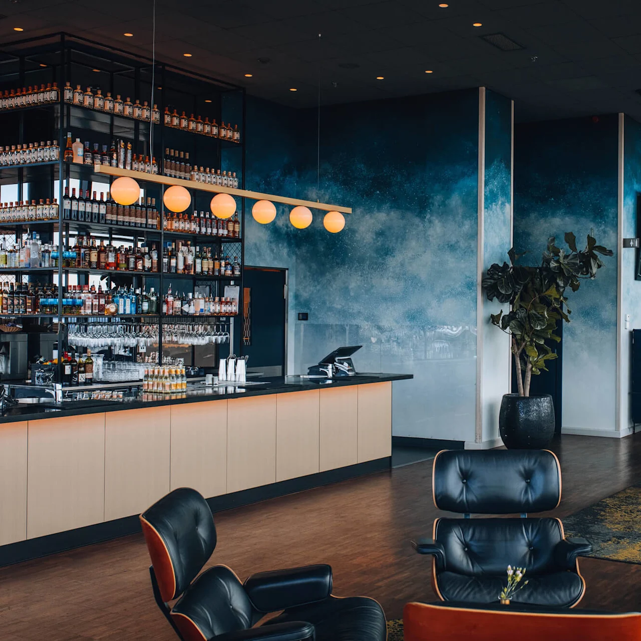 En moderne bar med elegante hylder fyldt med flasker, under sfæriske lys; omgivet af blå komfortable siddepladser og dekorative planter, hvilket skaber en luksuriøs loungeatmosfære.