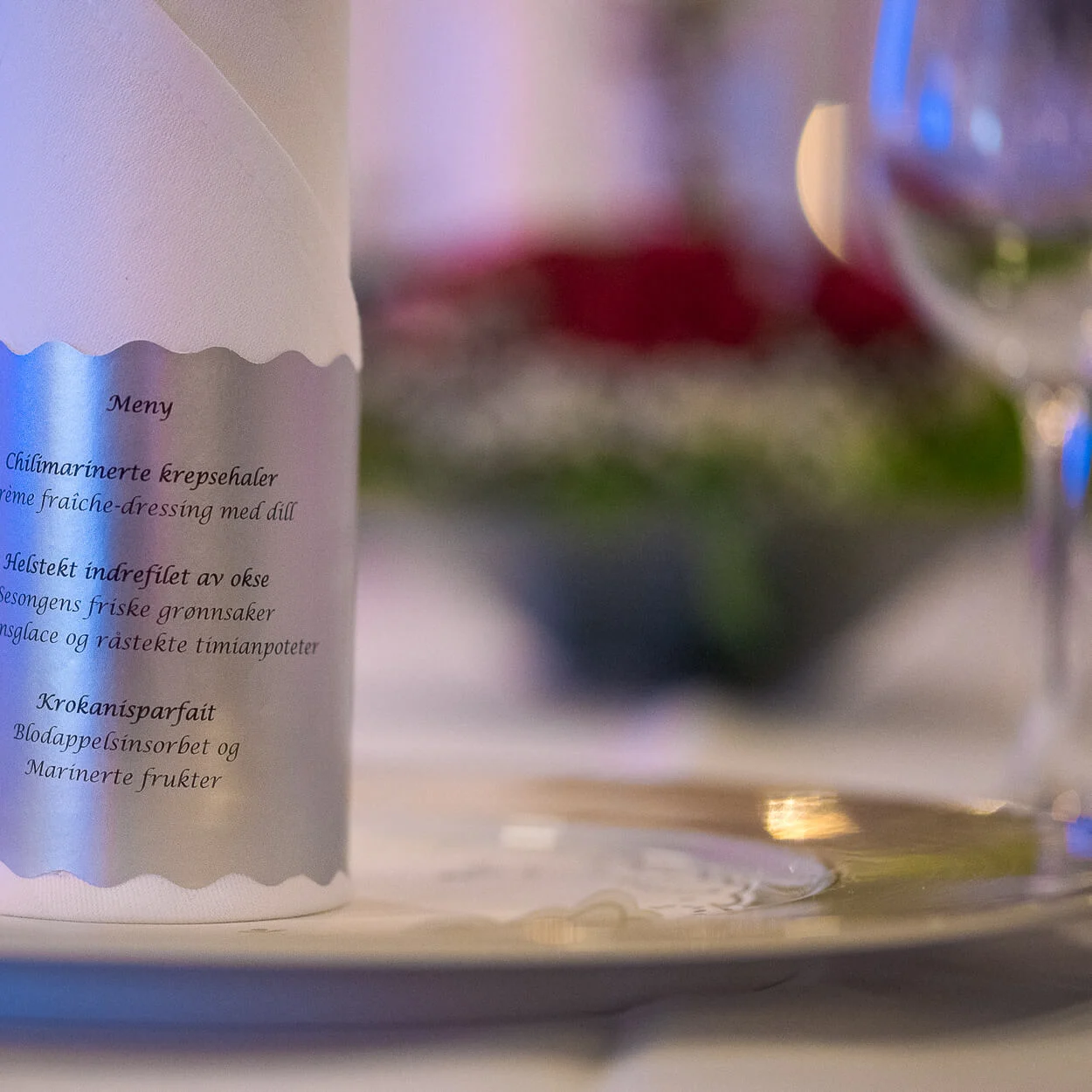 En rullet menu står på en dekorativ tallerken, og viser elegant retterne: "Meny, Chilimarinerte krepsehaler, Helstekt indrefilet av okse, Sesongens friske grønnsaker, Krokanisparfait."
