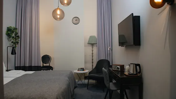 Bädden ligger under moderna hänglampor i ett hotellrum, bredvid ett litet skrivbord med en vattenkokare, stol och väggmonterad TV. Grå gardiner och subtil inredning skapar en mysig atmosfär.