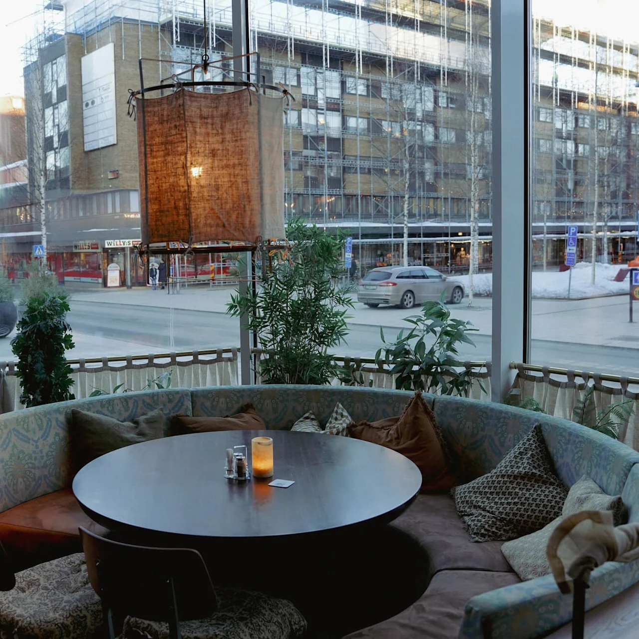 Hyggeligt caféhjørne med rundt bord, sofa og puder, udsigt til en bygade med bygninger og biler.