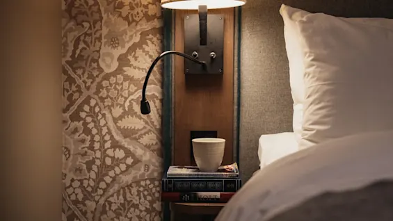 En mysig sängplats med en lampa, en hög böcker inklusive en med titeln HOLT, och en kopp, bredvid en bekväm säng.