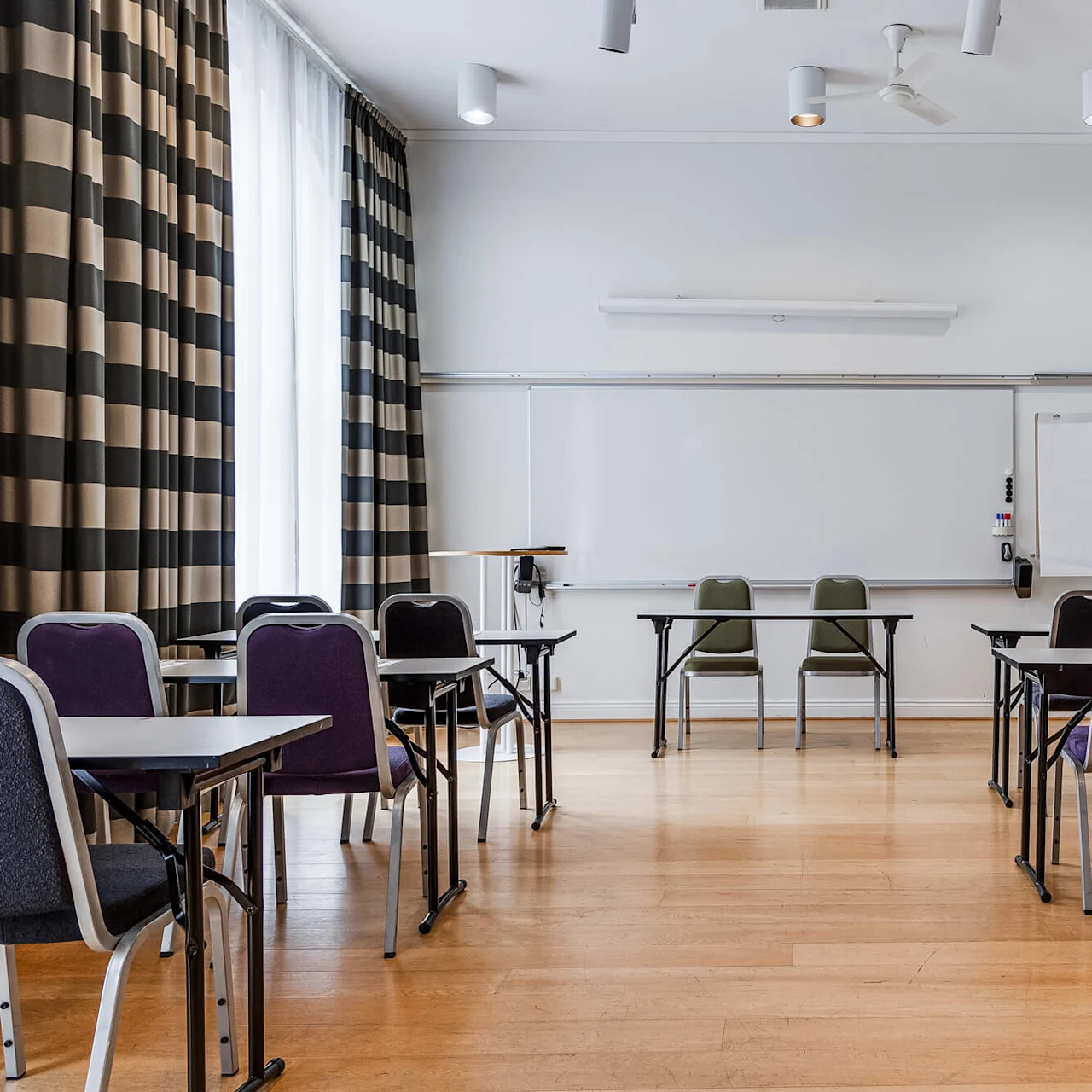 An empty classroom with desks and chairs arranged in rows, facing a whiteboard and projector screen on the front wall.