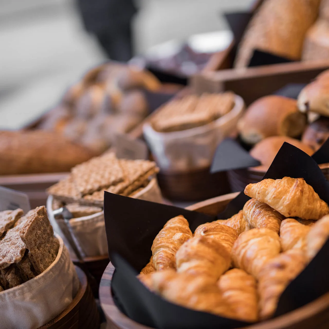 Croissanter ligger i en trekorg med svart papir, omgitt av andre brød og knekkebrød på et bord; et lite skilt merket "CROISSANT" er synlig.