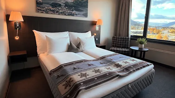 Ljust och mysigt hotellrum med en bäddad dubbelsäng, mönstrat överkast och utsikt mot fjäll genom fönstret.