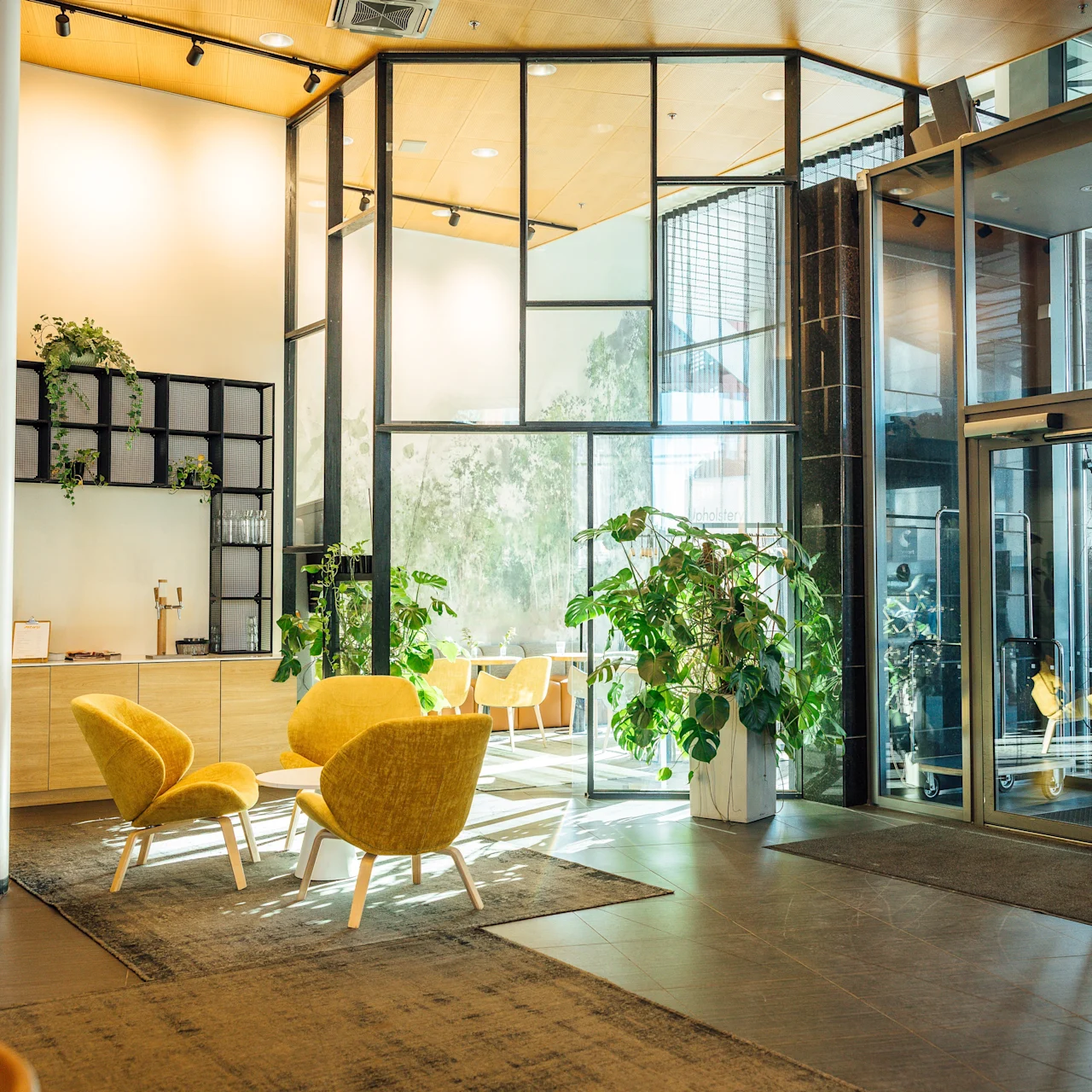 Bright lounge with yellow chairs surrounding a low table, positioned near large glass doors. Indoor plants decorate the space, adding a natural touch. Wooden and metal accents complement the modern design.