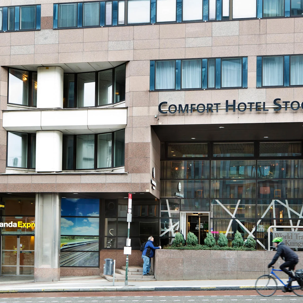Fodgængere og cyklister passerer forbi indgangen til Comfort Hotel Stockholm i et urbant miljø med skiltet "World Trade Center Kungsbron 1".