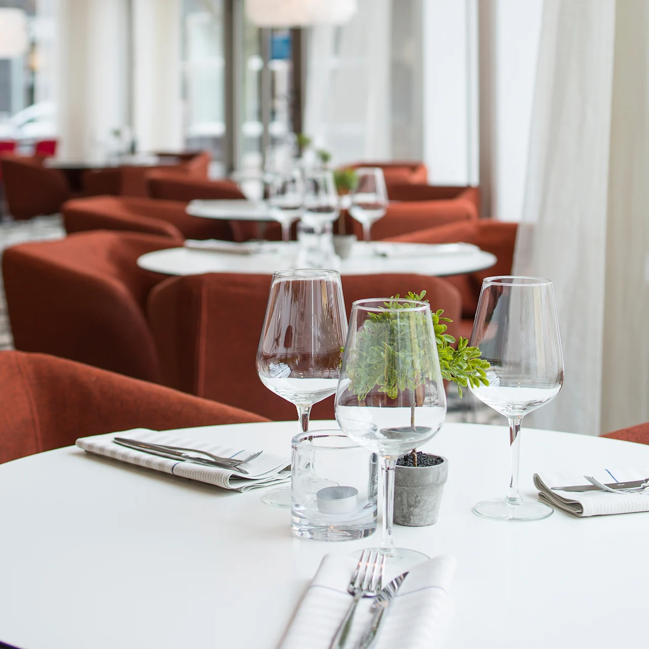 Rundt hvitt bord med tomme vinglass, potteplante og serviettinpakket bestikk i en moderne restaurant med røde lenestoler og store vinduer.