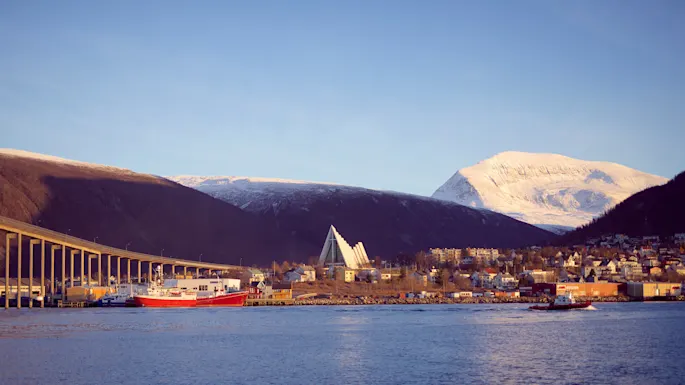 A coastal townscape with a bridge, boats, and a prominent triangular building against snow-covered mountains at sunset.