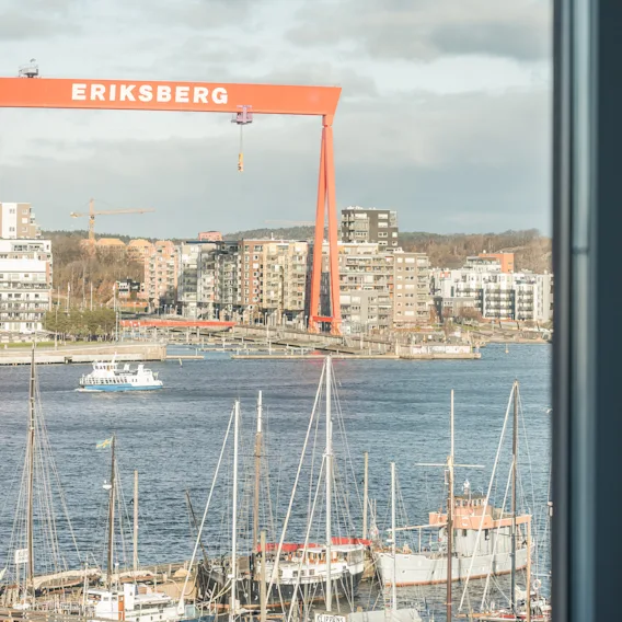 Utsikt från ett fönster över en hamn med båtar, en stadssilhuett och en stor röd kran märkt ERIKSBERG.