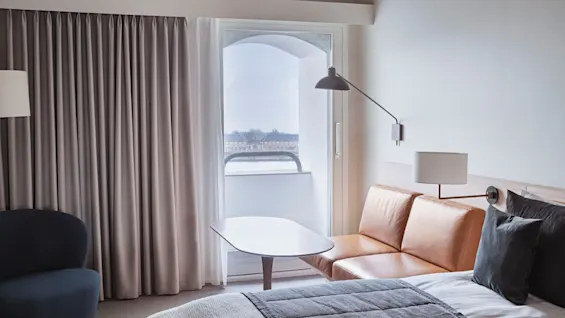 A cozy hotel room features a brown leather sofa near a window with sheer curtains. A small lamp hangs above, and a neatly made bed is visible. A soft armchair sits nearby.