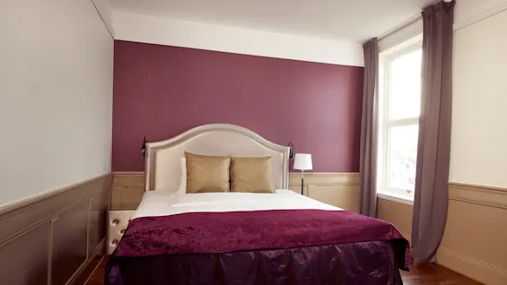 En pent oppredd seng med et hvitt og lilla tema mot en maroon vegg. Rommet er lyst med naturlig lys fra et vindu.