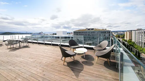 Rooftop terrace with modern chairs and tables arranged on a wooden deck. Surrounded by glass railings, it overlooks a cityscape with buildings and water under a partly cloudy sky.